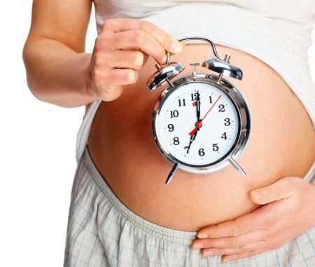 Closeup of a pregnant woman holding an alarm clock at her belly, focus on clock